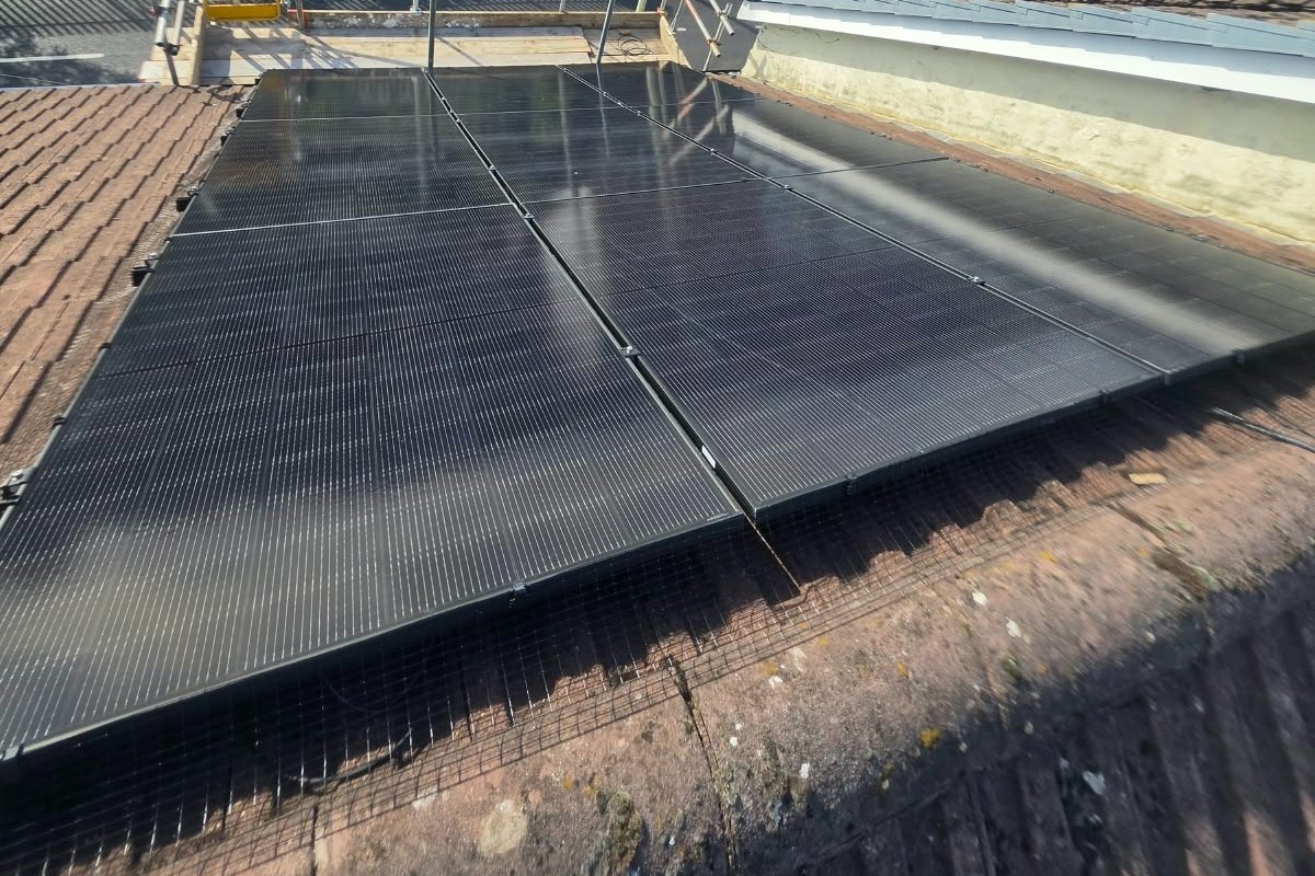 A solar panel array on a tiled roof beside scaffolding, with houses on a wooded hillside behind, photographed by Cozy Home Solutions.