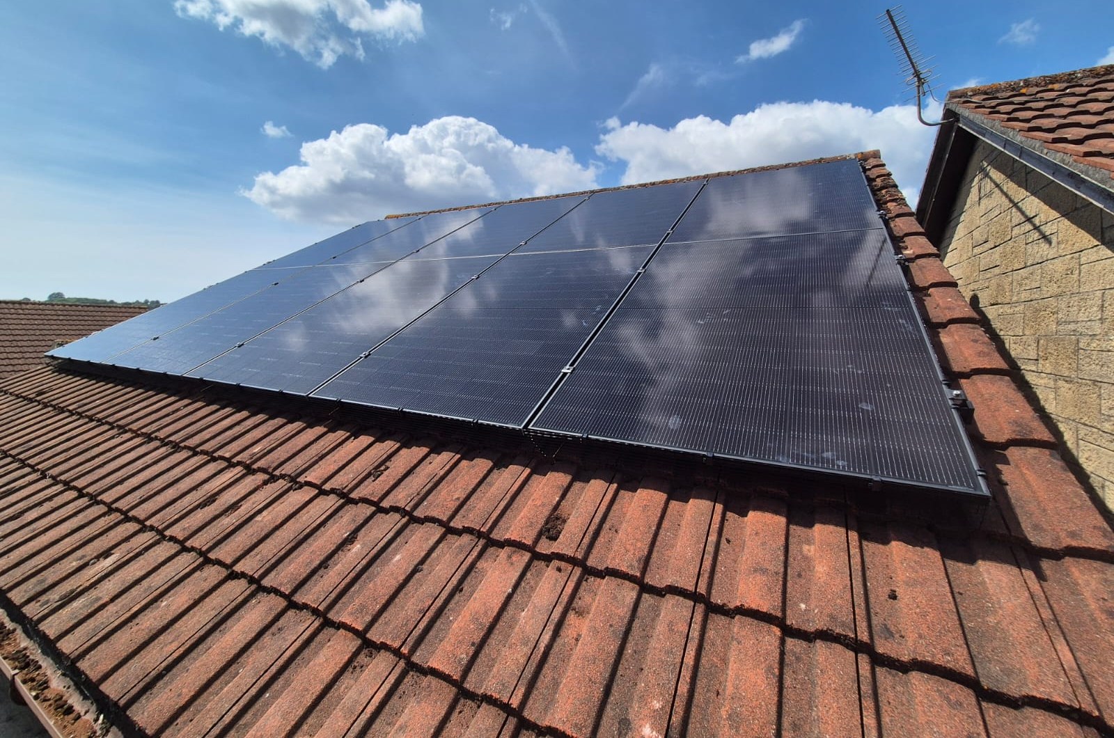 All-black solar panels covering a red-tiled roof slope beside a brick chimney, photographed from the scaffold at a Newport installation.