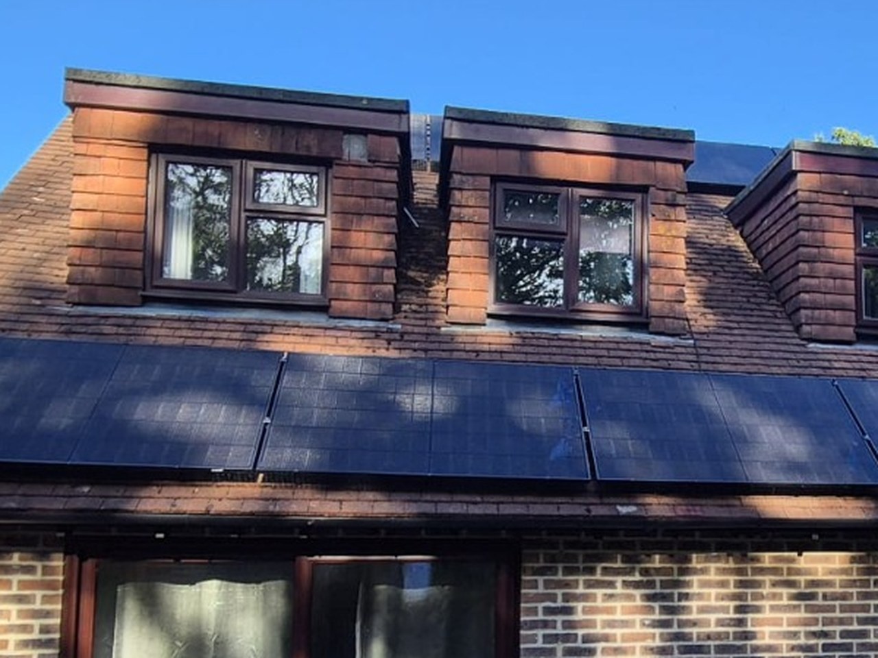 A row of five all-black solar panels fitted below three dormer windows on a brick home's tiled roof, photographed by Cozy Home Solutions.