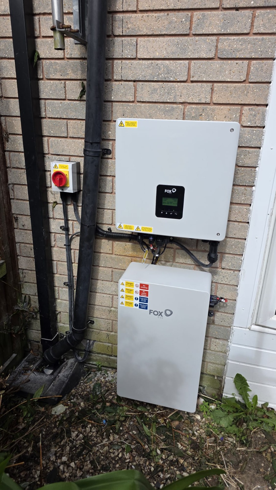 Fox ESS inverter with a red isolator switch and a Fox ESS battery unit mounted on an exterior brick wall.