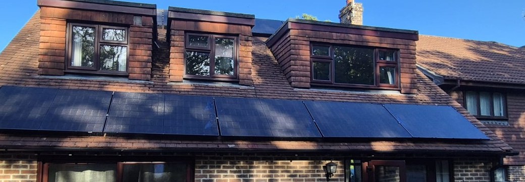 Solar panels mounted along the lower tiled roof slope of a brick home beneath three dormer windows, photographed by Cozy Home Solutions.