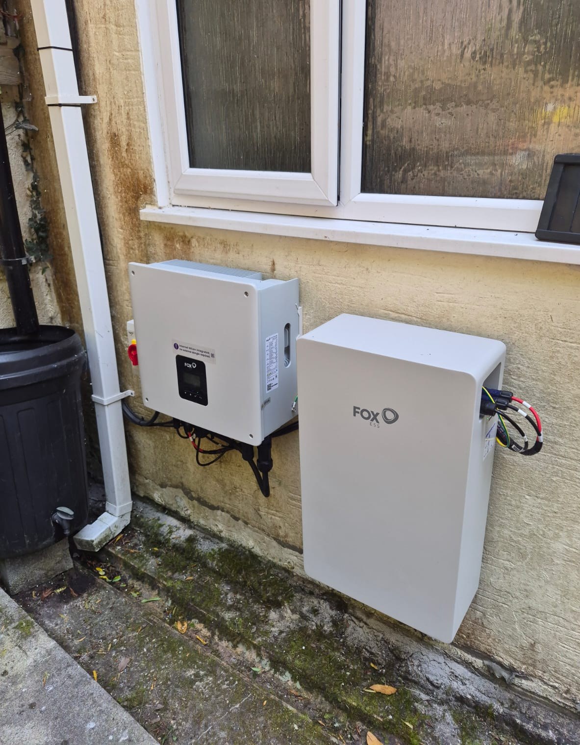 Fox ESS inverter and a white wall-mounted battery unit fixed to the exterior wall of a house beneath a window.