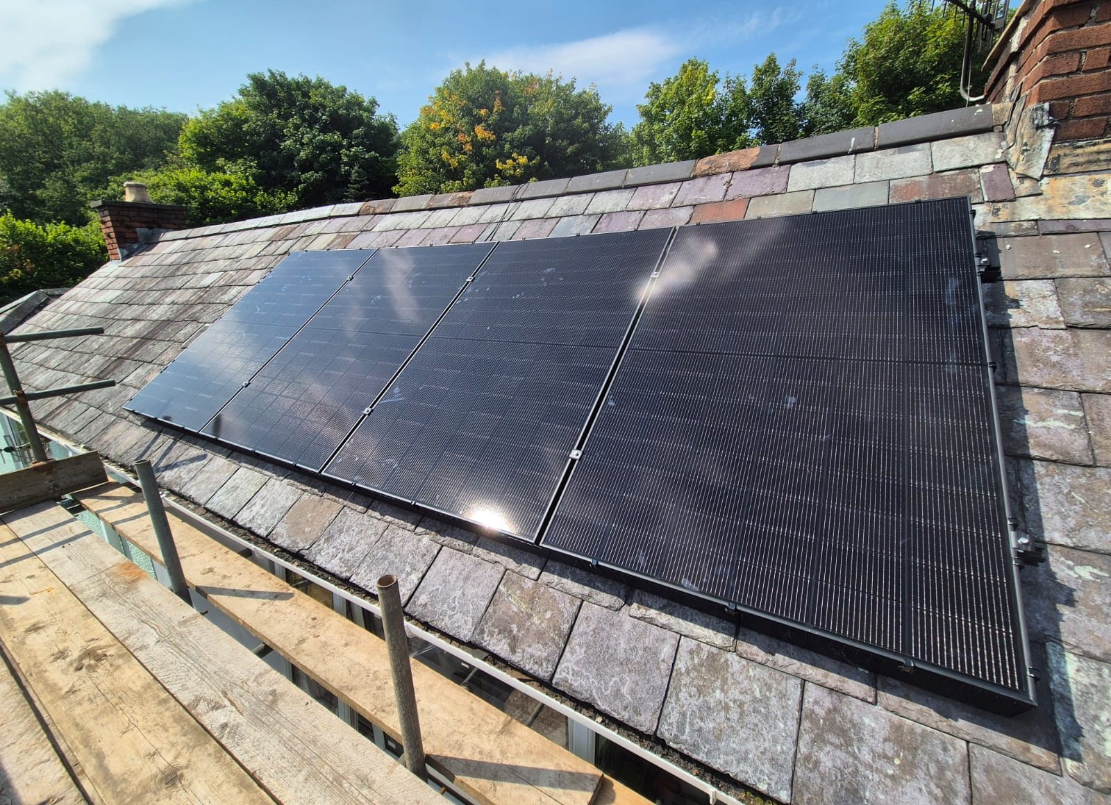 All-black solar panels fitted to a slate pitched roof beside a brick chimney, seen from scaffolding during a Cozy Home Solutions installation in Swansea.